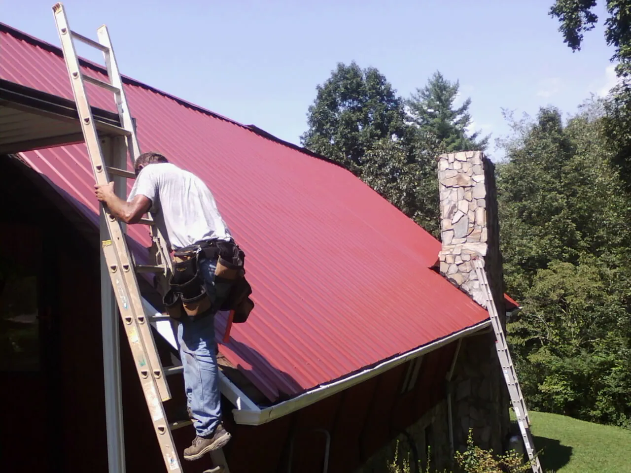 Roofer on a ladder beside a red metal roof and stone chimney — Soffit & Fascia Repair by Roofing Brothers in Argusville